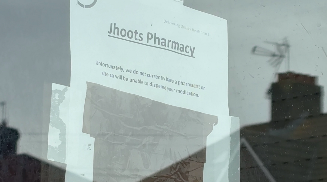 An A4 sheet of paper is taped to the inside of a glass door. It reads 'Jhoots Pharmacy' in big, bold and underlined letters at the top. In smaller type, it then says "Unfortunately, we do not currently have a pharmacist on site so will be unable to dispense your medication."