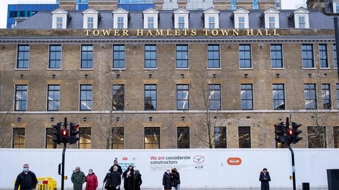 A picture of Tower Hamlets town hall, a brown brick building with white hoardings along the ground floor. The view shows people waiting at a set of traffic lights ready to cross, and in the foreground there is a road,