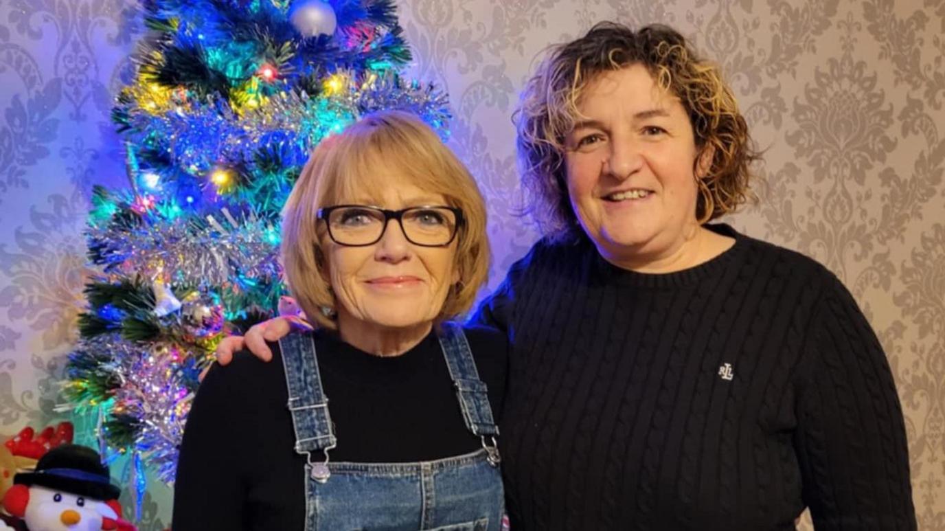 Gail and Catrina Gourlay smile at the camera while standing in front of a Christmas tree. Gail wears blue denim dungarees and a black top and Catrina wears a black jumper.