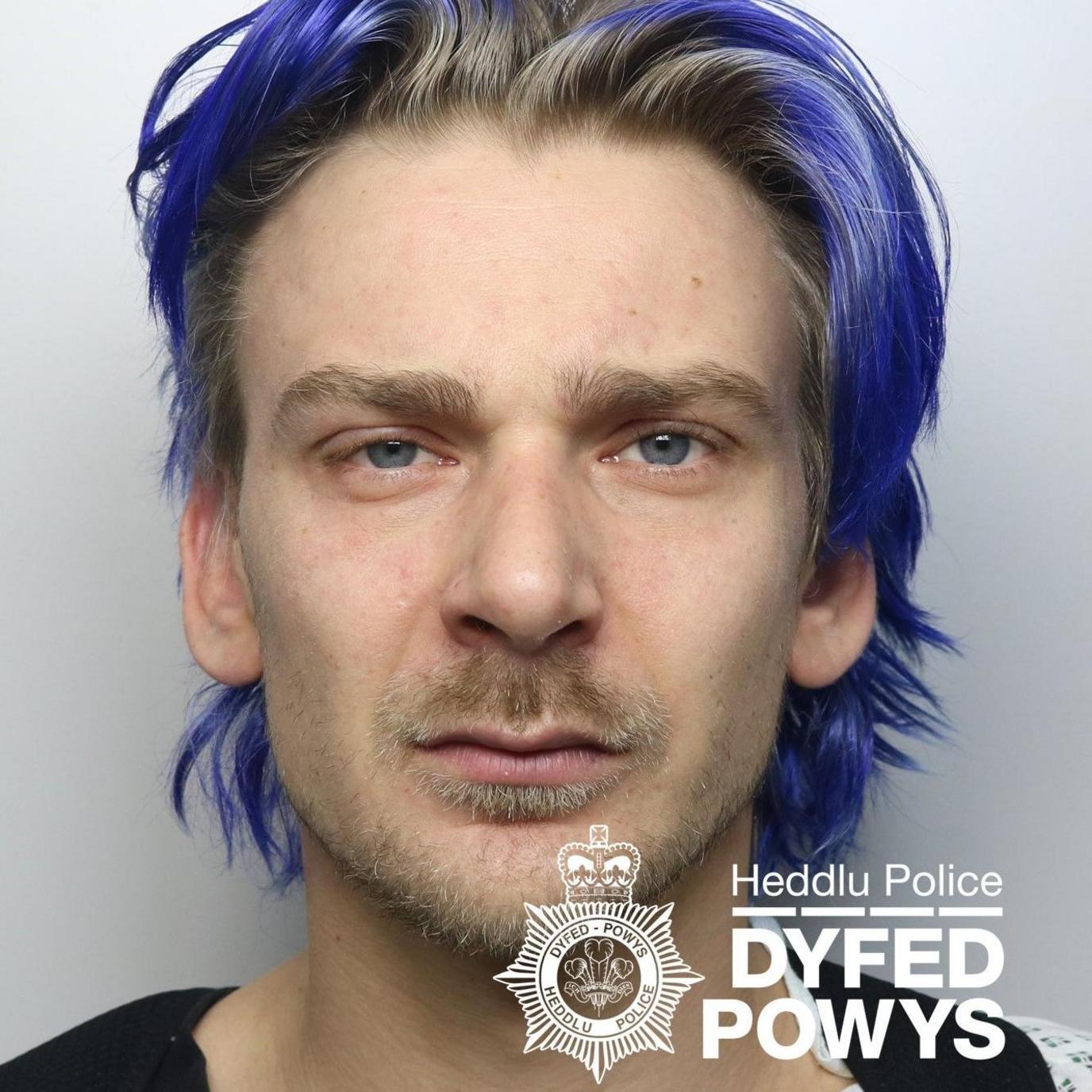 Mateusz Sikorski, in a Dyfed-Powys Police mugshot. He is staring at the camera, and has mostly dark hair, with blue streaks