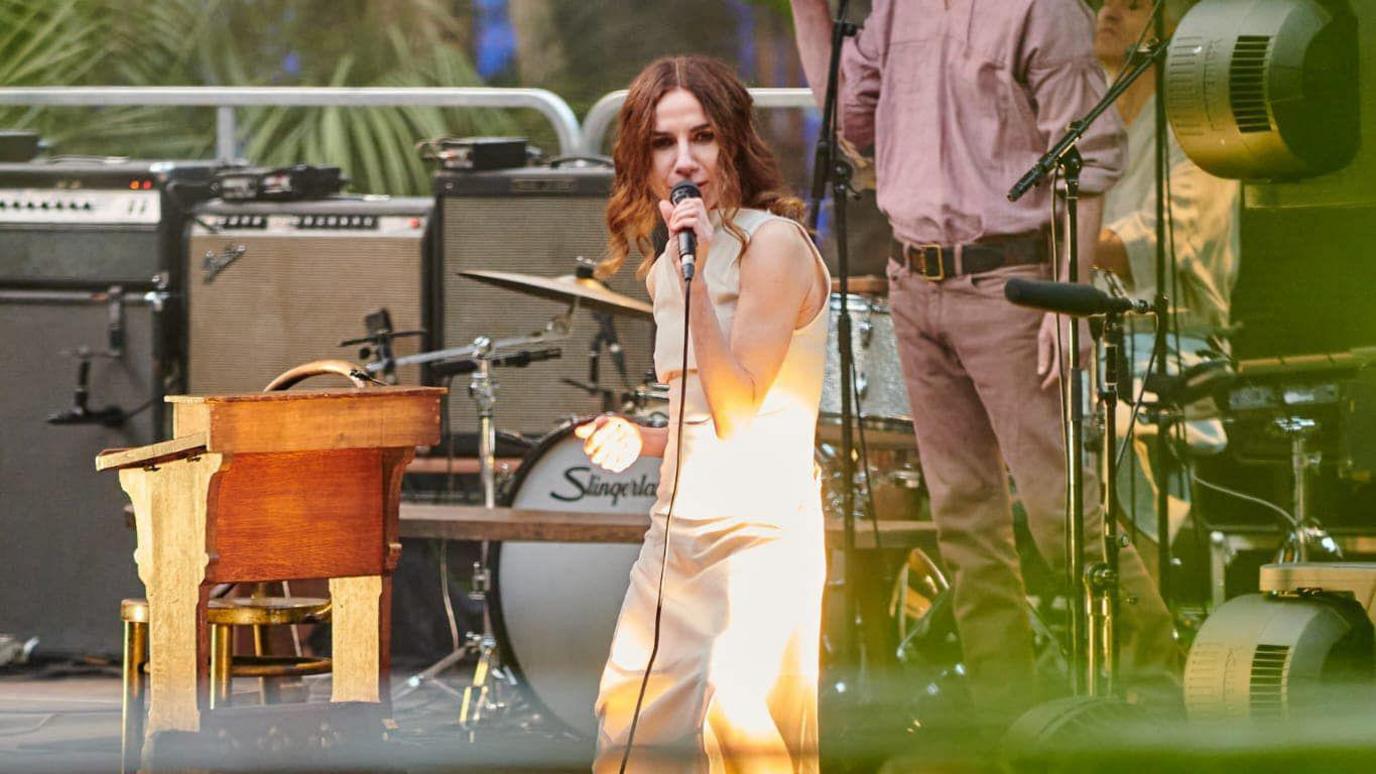 PJ Harvey singing into a microphone. She has shoulder length curly brown hair and is wearing a fitted white dress.