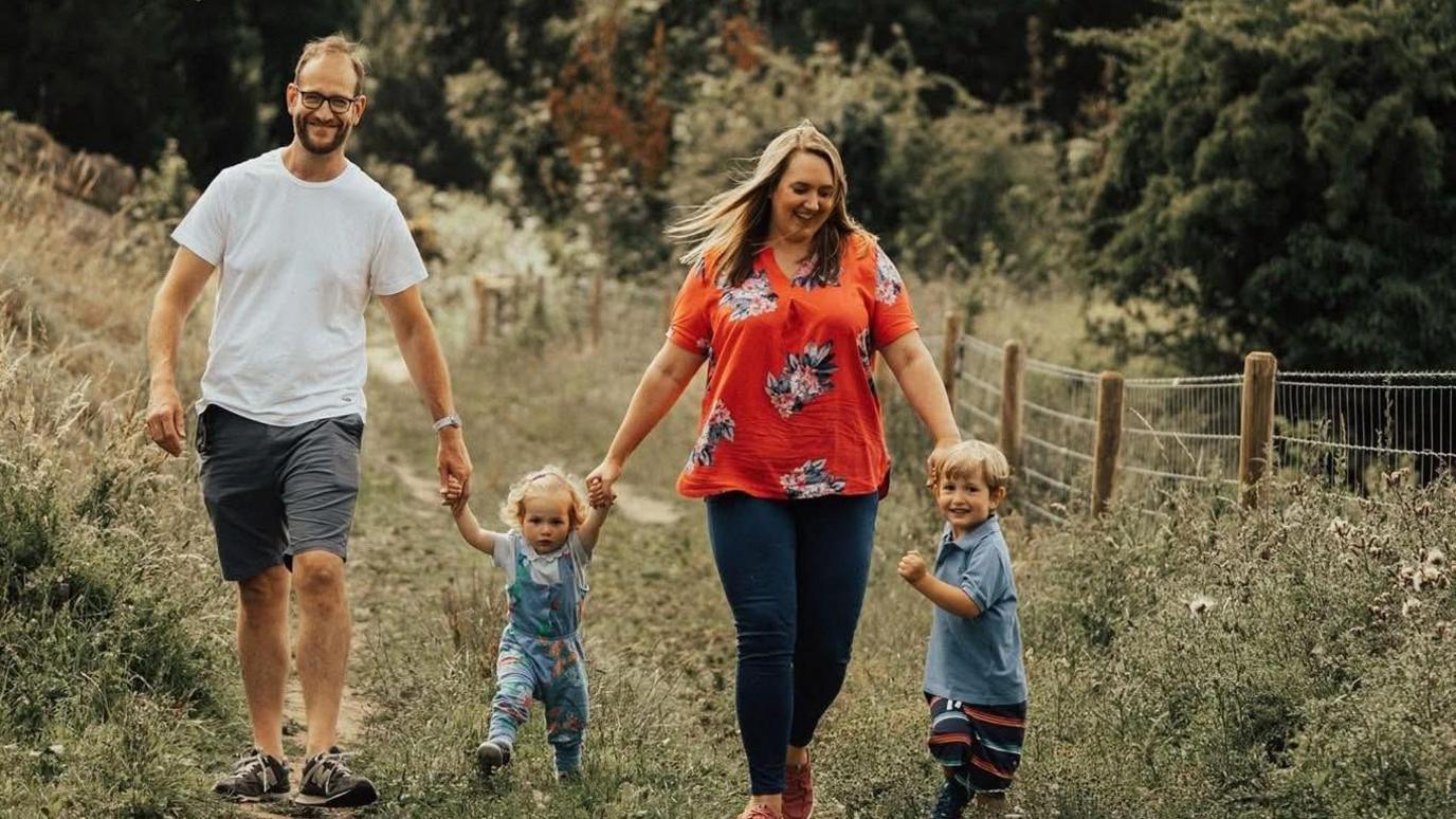A man and woman holding the hands of their two children, a young girl and boy, walking along a grass path. The man is wearing a white top and grey shorts with trainers and glasses and smiling at the camera. A little girl is wearing blue dungarees. A woman is looking down at her little boy and has long blonde hair and an orange flowery top and blue jeans. The little boy is standing to the side and smiling at the camera.