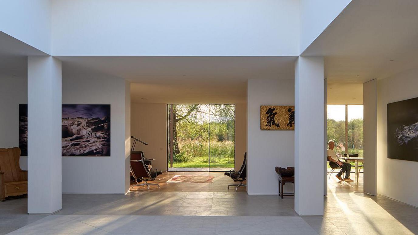 Modern building with white walls. Two pillars hold up the white roof. There are pictures hanging on the wall behind, and glass doors beyond reveal a green garden with a tree behind. A person is sitting at a table in a room to the right.