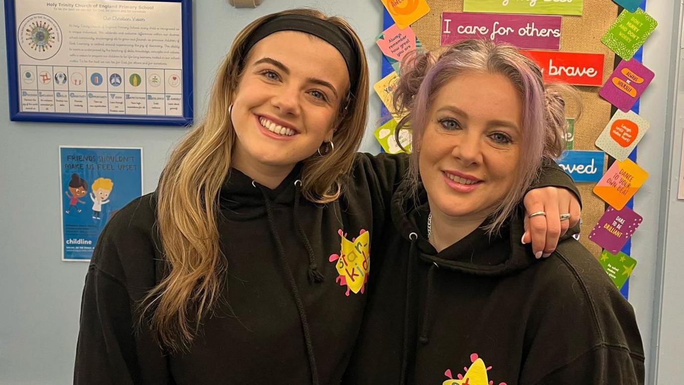 Ellie and Jan Hayes smile at the camera. They are both wearing black hoodies with a yellow and pink Starkidz logo on them. They are in a classroom.