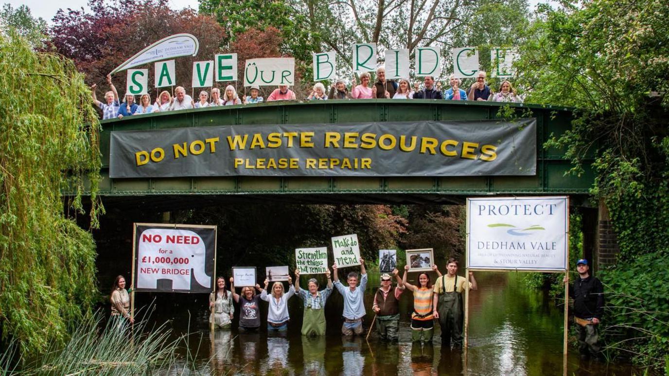 People standing on a bridge and in the water holding protest signs and surrounded by greenery.