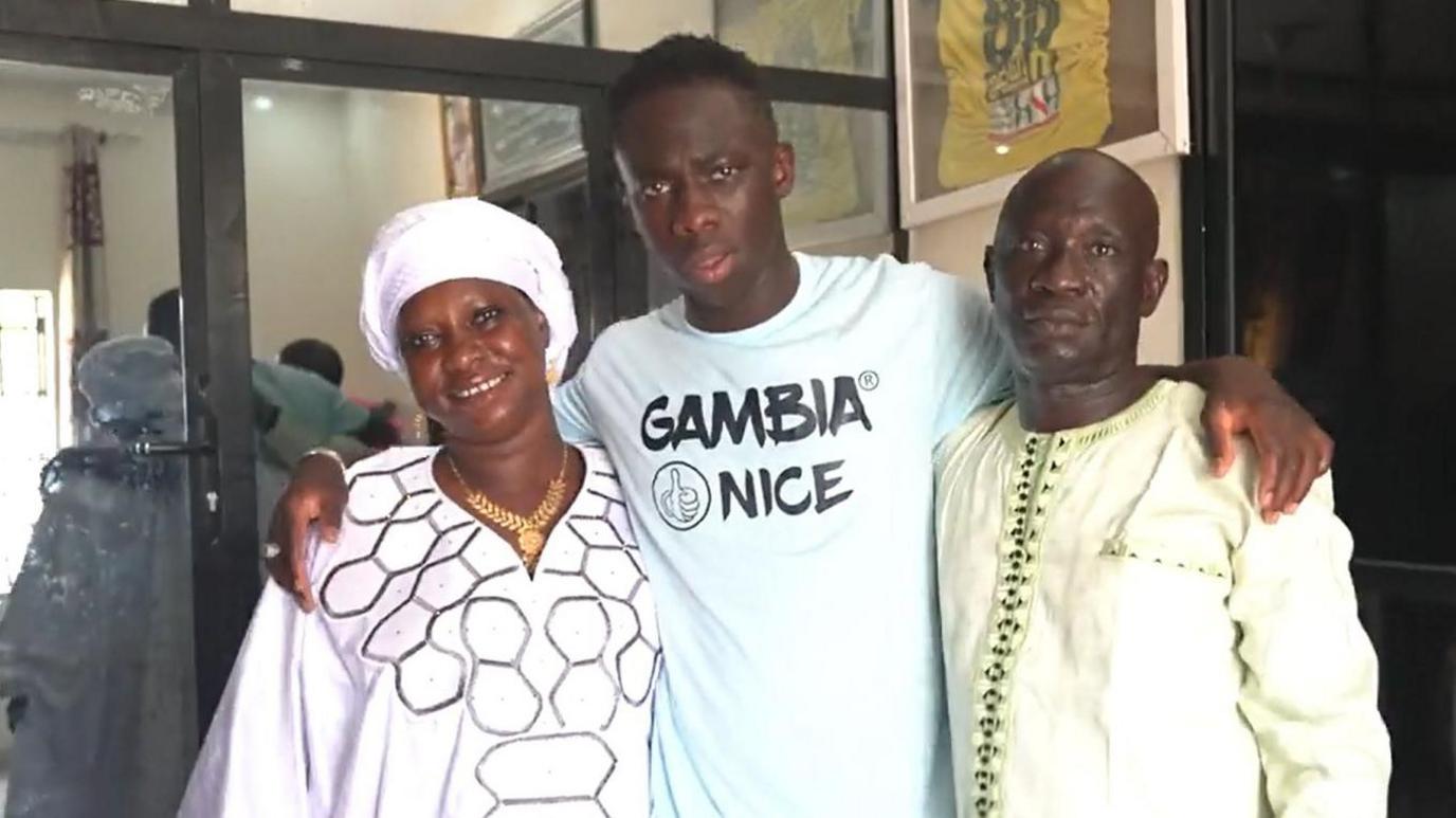Yankuba Minteh, wearing a T-shirt bearing the message 'Gambia Nice' and a thumbs up cartoon image, puts his arms around his mother Jassey and father Buhari, who stand facing the camera on the left and right of their son respectively. Jassey wears a white head covering, gold necklace and traditional white robe, while Buhari is in a light green coloured robe.