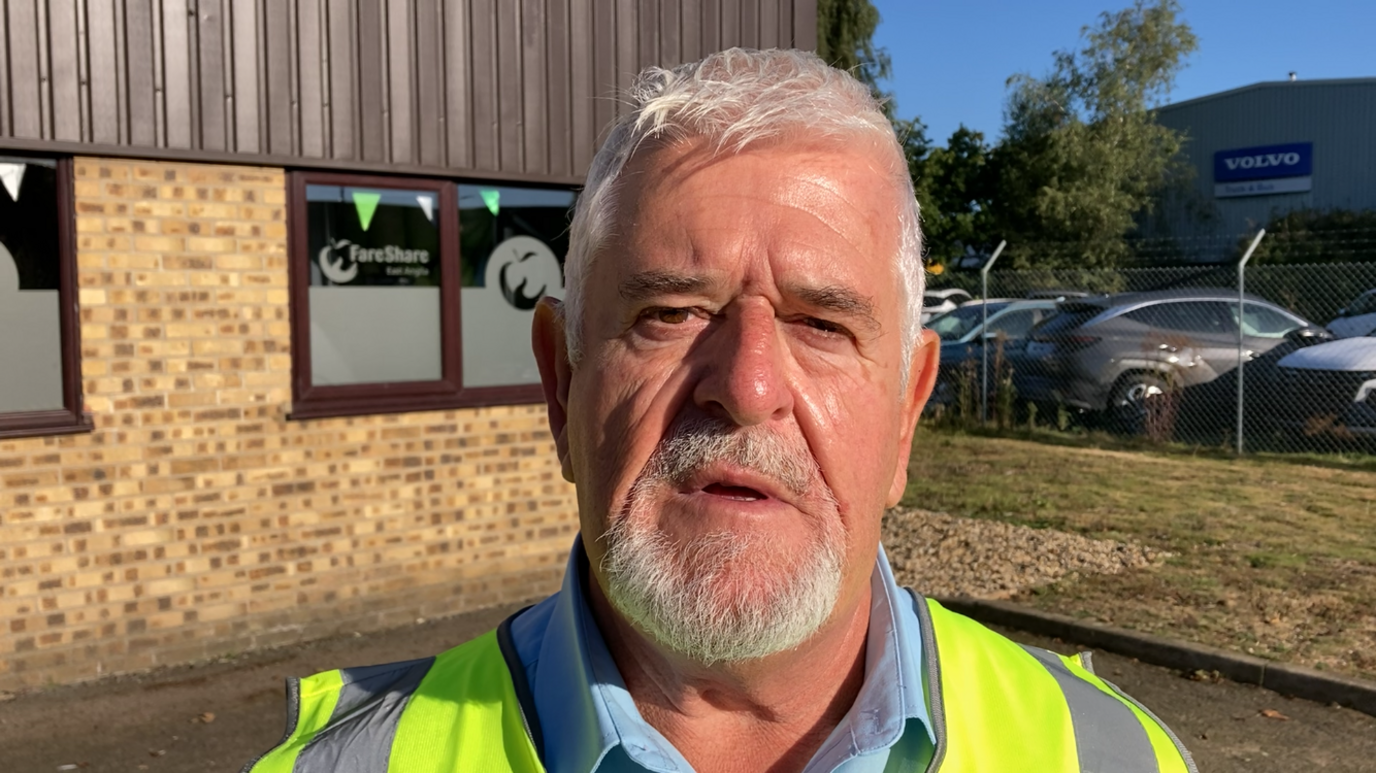 Mike Barrett wearing a hi-vis vest over a blue shirt. He has short grey hair and a goatee. He has his mouth slightly open and is standing in front of a brick building.