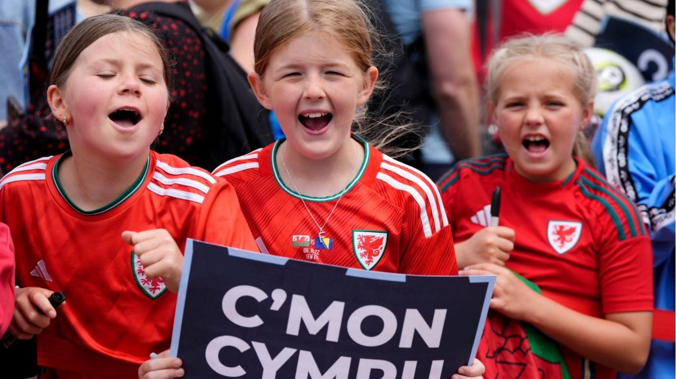 Three young girls all wear red Wales shirts and are cheering as the girl in the middle holds a sign with the words C'mon Cymru