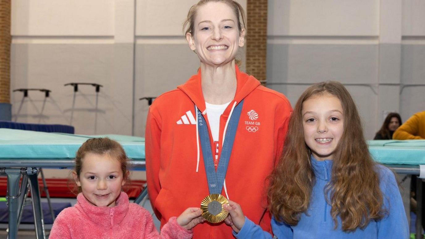 Page stands proudly showing off her medal, which is being held by two young girls