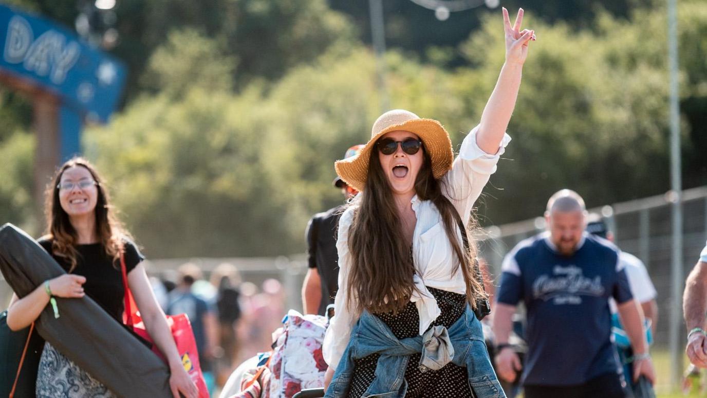 A woman wearing a straw hat and making the peace sign as she arrives at Isle of Wight Festival