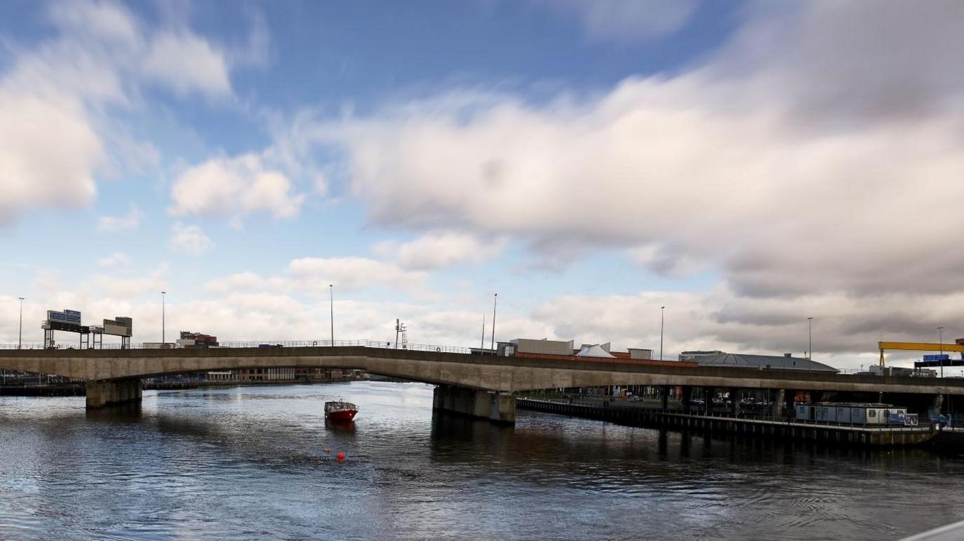 A wide shot of the Lagan Bridge in Belfast. It's a large concrete bridge over the Lagan River. In the distance is one of the yellow Harland and Wolff cranes, and the top of the SSE Arena and Odyssey pavillion buildings. It is a cloudy day, with some breaks of blue sky.