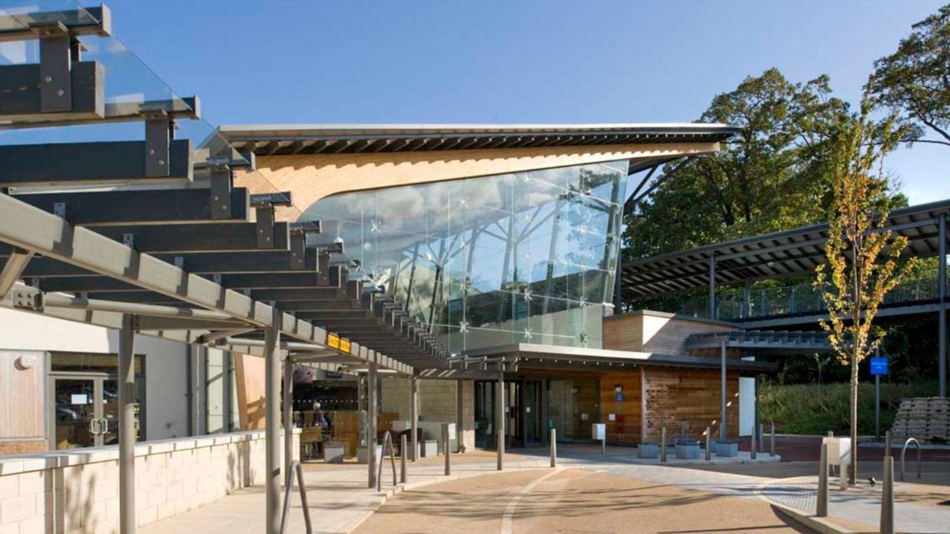 The entrance to a hospital with a road and path leading up to it. The main building has a slanted ceiling and a large open window either side with two longer buildings either side of it.