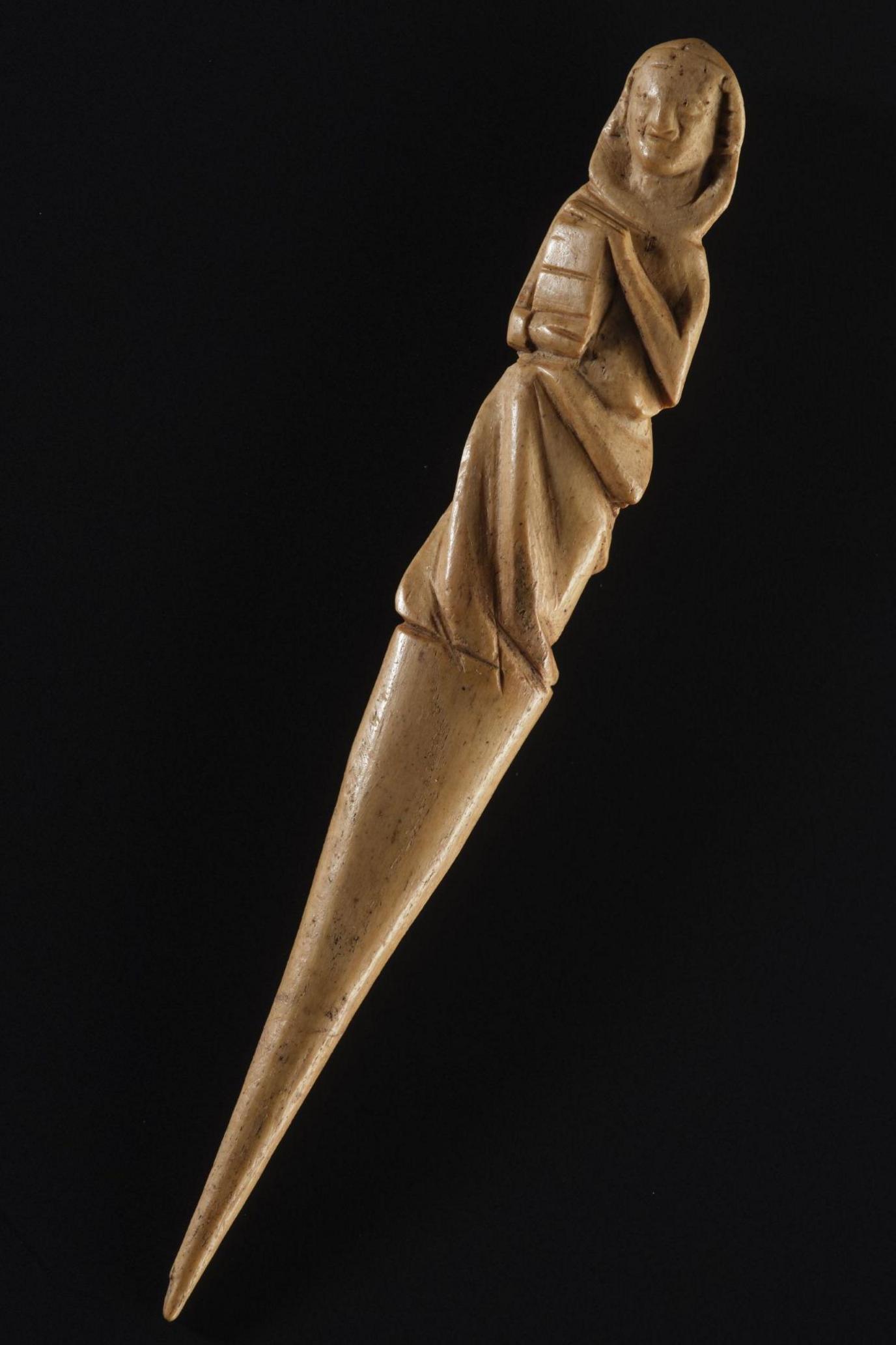 A close up of the gravoir, a needle-shaped ornately carved piece of red deer antler. The carving is of what looks like a hooded figure holding a book. The object is photographed against a black background.