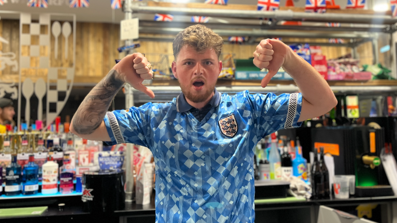 Barty Williams, wearing a blue England polo shirt, he has a disapproving expression and is holding both his thumbs down, he is standing at a bar