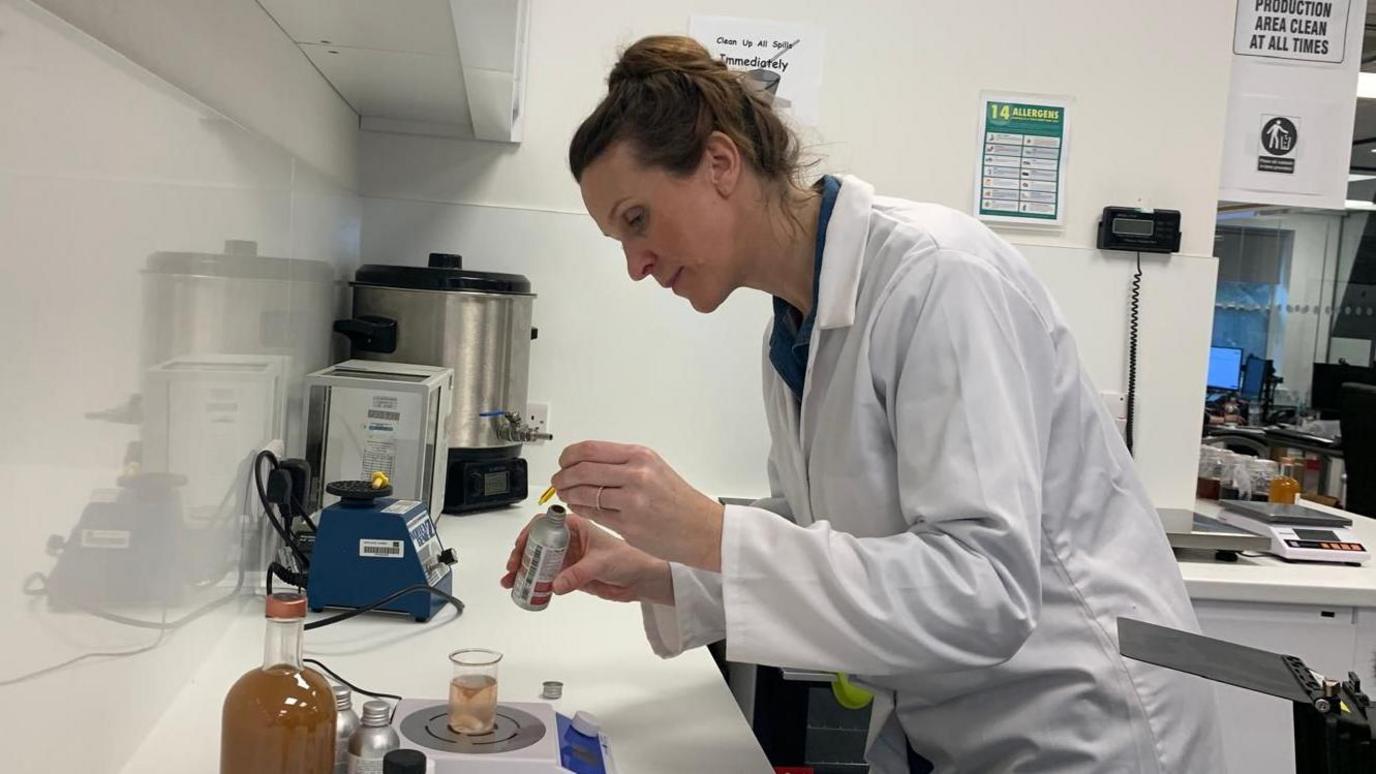 A woman in a lab coat uses a pipette to drop a liquid into a beaker. She stands in a research and development lab.