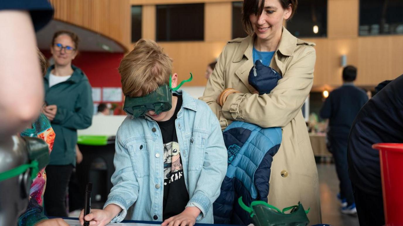 A small boy is drawing while wearing protective goggles and a blue shirt. He is watched by a dark-haired woman in a light brown coat.