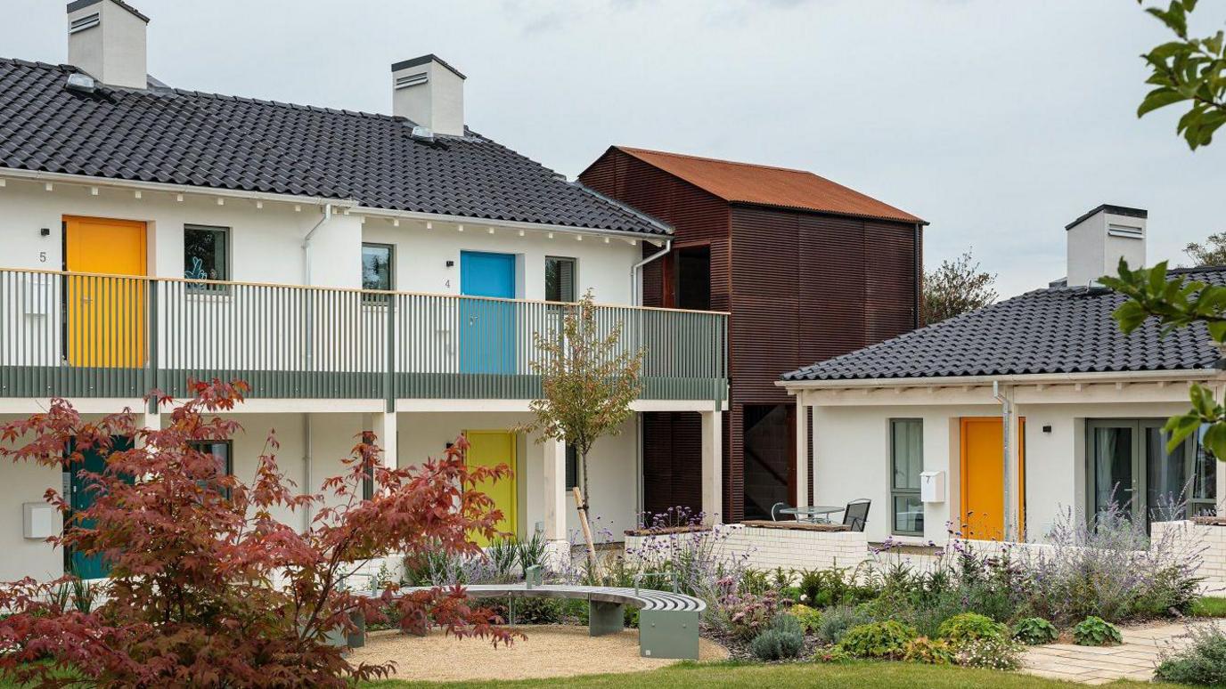 Two-storey building to the left and one-storey building to the right. THey have grey roofs with small white chimneys. The buildings are divided into different flats with coloured doors and a railing in front on the two-storey structure. There are shrubs and trees in the foreground and a semicircle seating area.