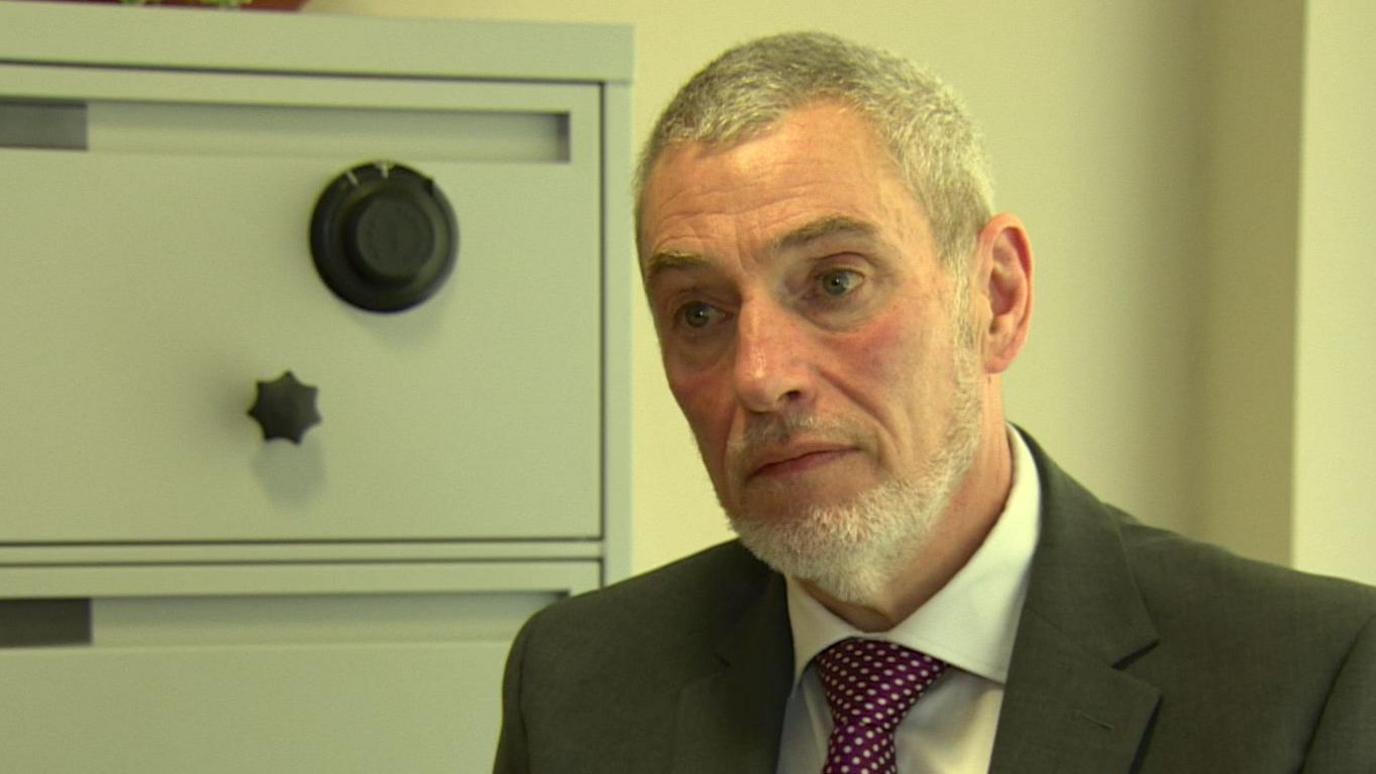 Hugh Hume. He has short grey hair wearing a dark grey blazer, white shirt and purple tie with with white polka dots. A filing cabinet is beside him