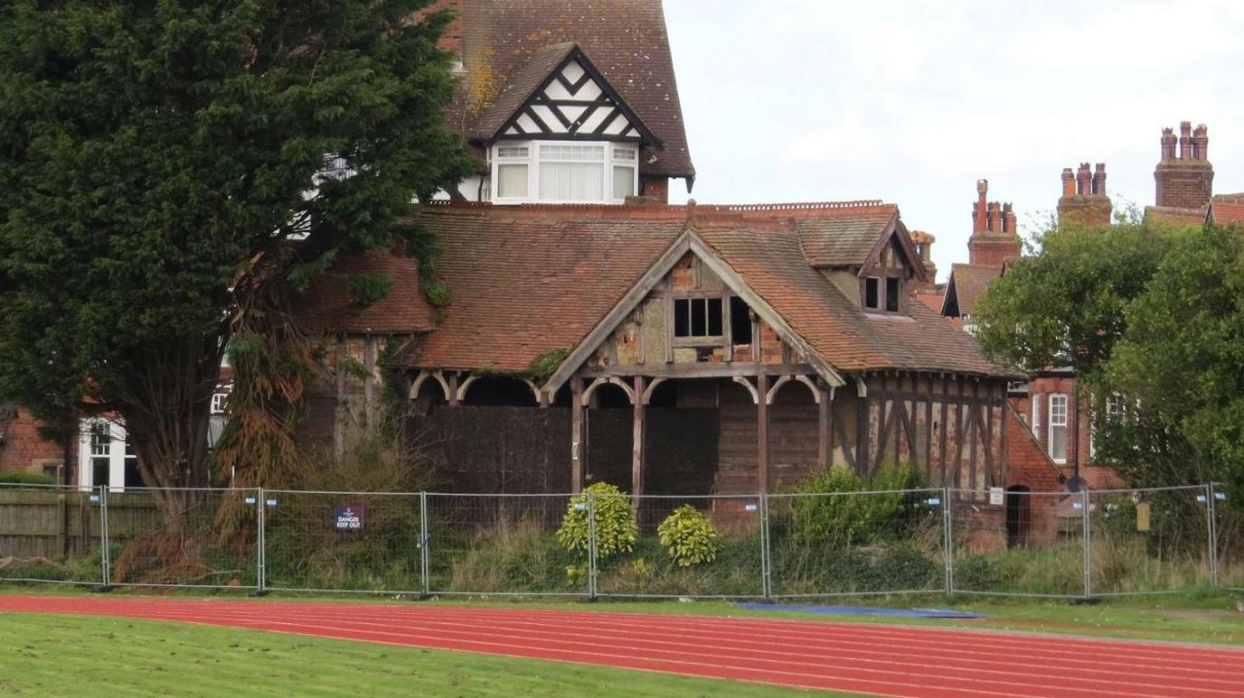 A dilapidated Tudor-style wood and brick building fenced off with metal fences on the side of an athletics track