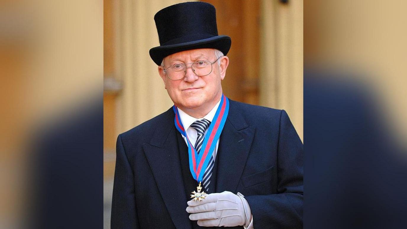 Oleg Gordievsky wearing a suit and top hat with a medal on a blue and red striped ribbon around his neck. The medal is gold in colour and has seven shapes protruding from its edges, one of which attaches it to the ribbon.