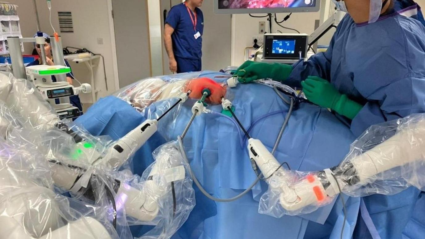 Robotic arms hold medical attachments against the body of a small baby, most of which is covered in surgical cloth and plastic wrapping. A surgeon wearing green gloves and blue scrubs can be seen holding a tool close to the baby body. More medical staff can be seen in the background along with a screen showing a close up of the baby. 