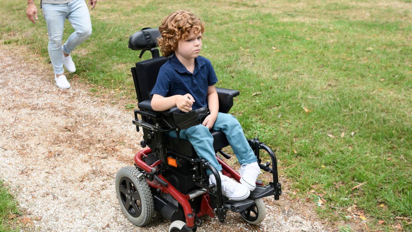 A boy around six or seven with curly hair in a red powered wheelchair on a path through grass.