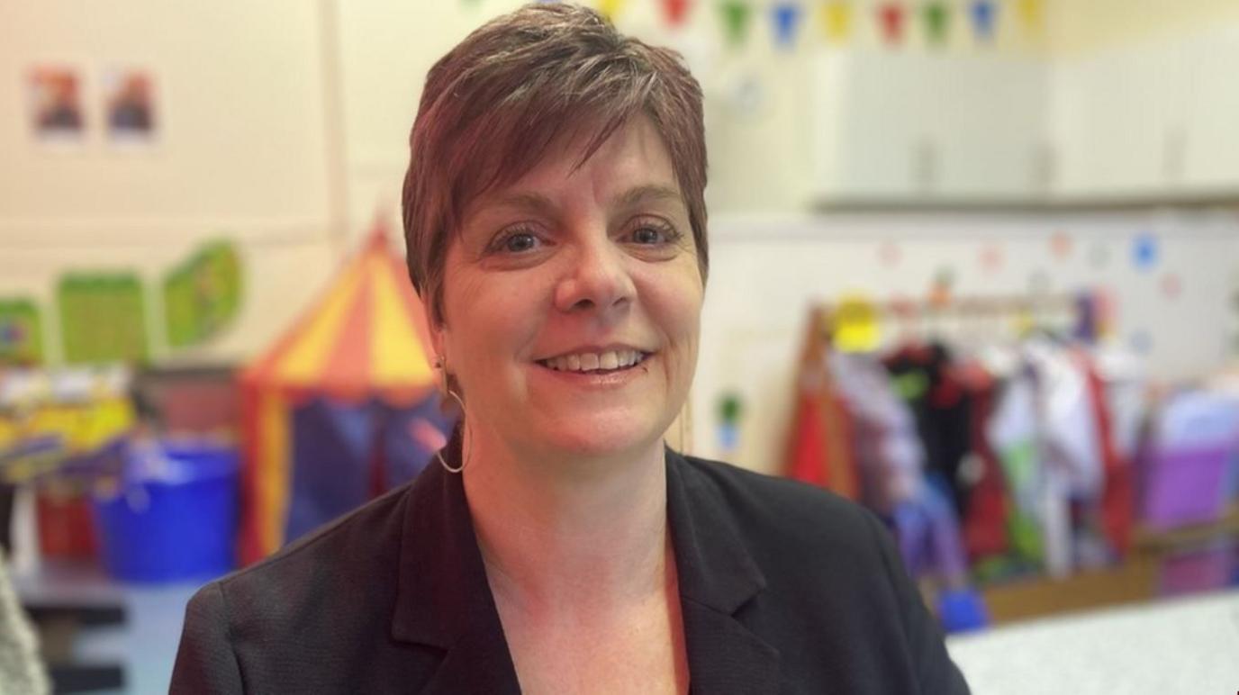A woman with short hair and wearing hoop earrings and a dark-coloured blazer standing in front of children's toys including a brightly coloured tent, bunting and a rail of dressing up clothes