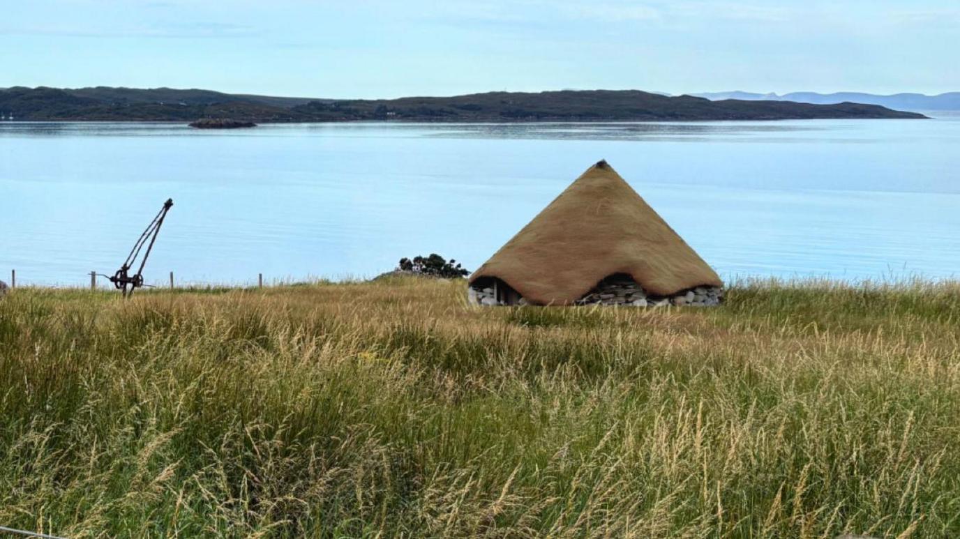 A visualisation of how the roundhouse would look at Gairloch Museum. The roundhouse has a conical, turf roof above stone walls. It is in an area of long grass and the sea is behind it.
