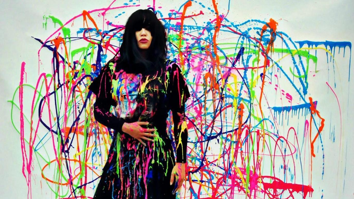 Y gantores Mared Lenny o flaen cefndir lliwgar gyda phaent wedi ei daflu ar ei hyd yn arddull yr artist Jackson Pollock