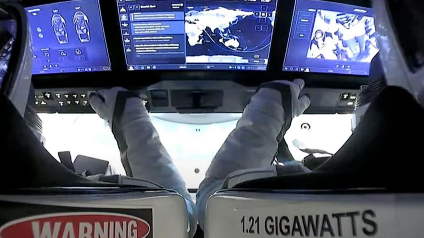 Two of the SpaceX crew carry out instrument checks during landing. They are facing the viewport and a bank of large toughscreens in the cockpit.