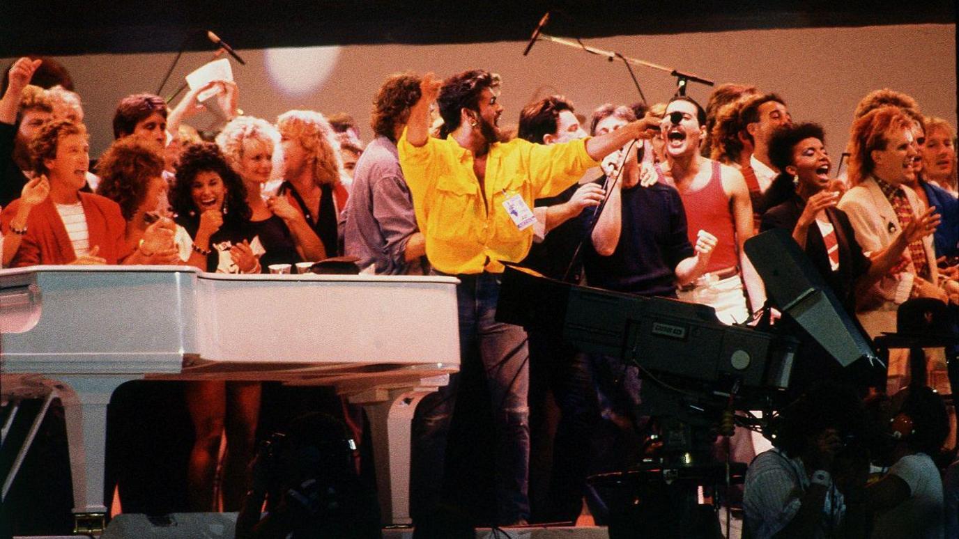 Musical stars gathered on the stage. At the front is George Michael in a yellow shirt holding the mic out.