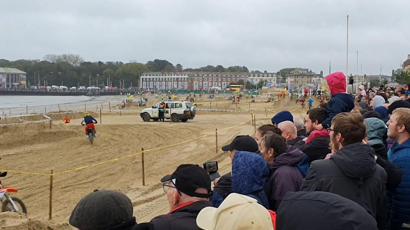 Spectators watching bikes from the side lines whizzing through the wet sand with buildings behind. It's cloudy.