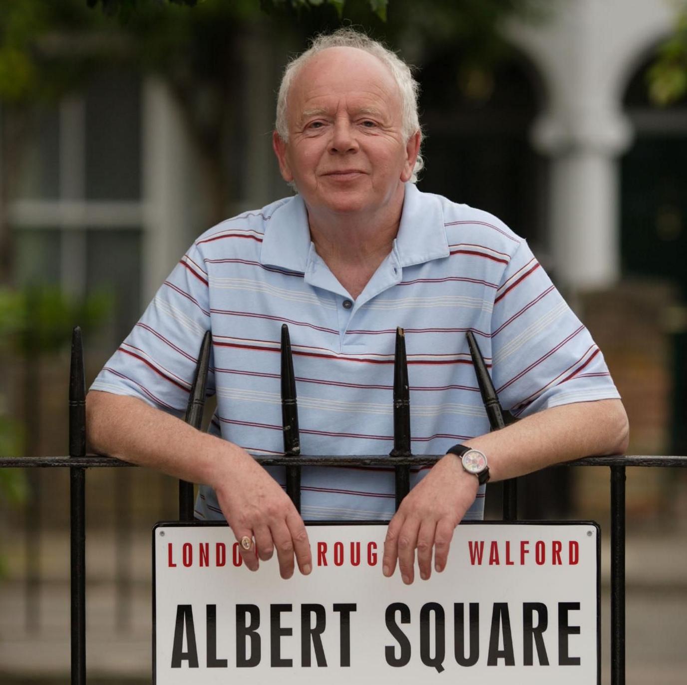 A posed photograph of Ray Brooks stood behind the 'Albert Square' sign on the set of EastEnders. He has short white hair and wears a striped red, white and blue polo shirt.