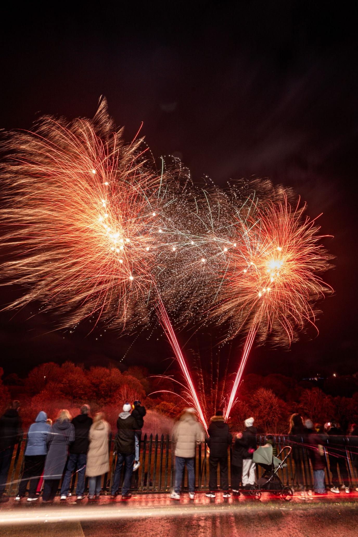 Brilliant red fireworks burst across a dark night sky, creating dazzling trails of light and sparks. A group of spectators stands in the foreground, watching the display from behind a railing.