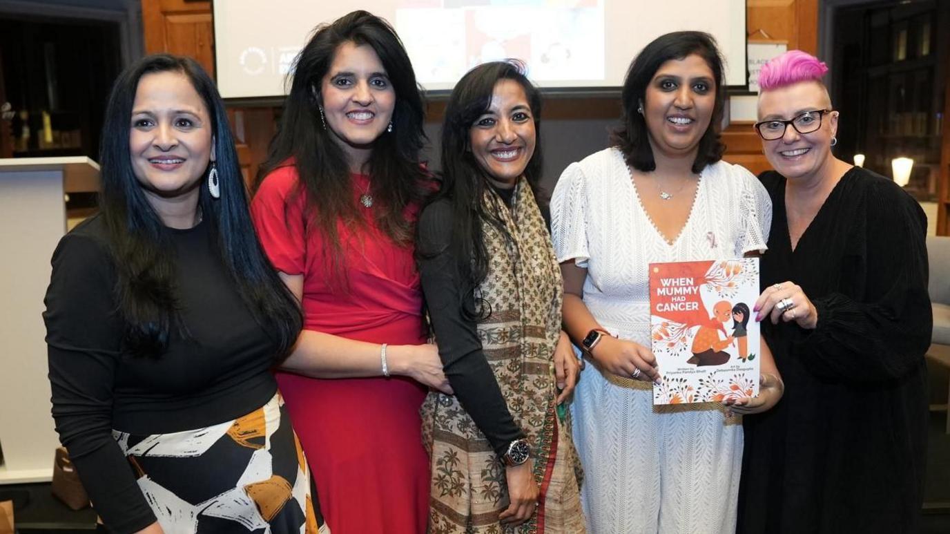 Five women standing in front of a screen, smiling and looking towards the camera. The woman on the far left is looking slightly away, with long dark hair, a skirt and black top, the next woman long is wearing a red dress, and has long dark hair, the woman in the middle has on a long silk scarf and dark top, with long dark hair, Priyanka, is wearing a white lace all in one trouser outfit, holding her book, and the woman on the right has short pink hair, is also holding the book and is wearing all black. 