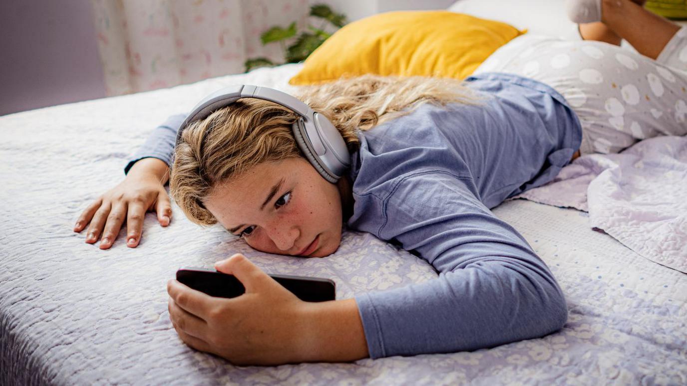 A young teenager lies on a bed with a phone in her hand and is wearing headphones.