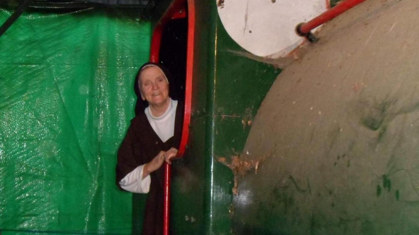 A green train engine cabin, appearing out of the cabin door is a nun from a local convent. There is a green tarpaulin sheet in the background.
