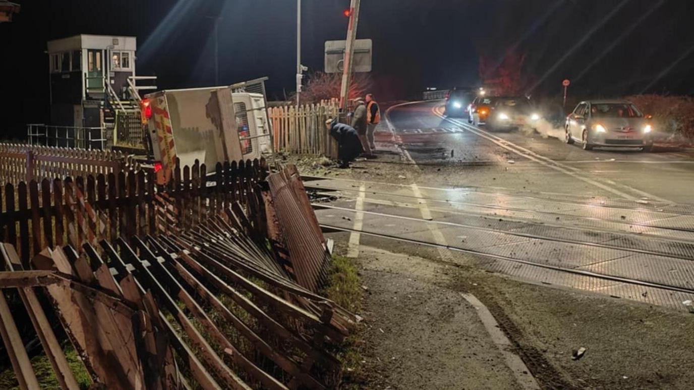 A damaged fence at a level crossing with a van on its side on rail tracks and a queue of traffic on a road