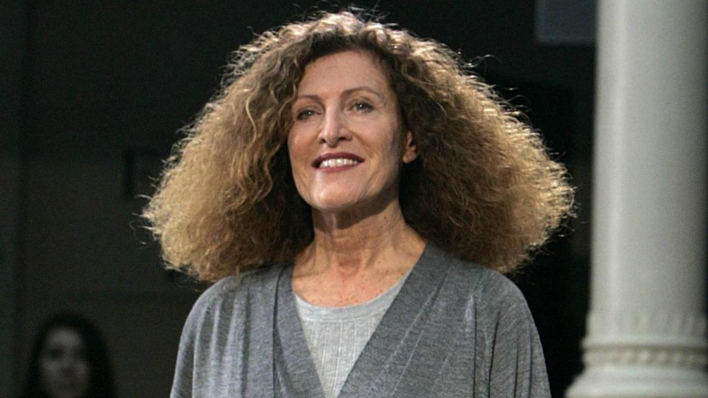 This is a photo of a woman with brown curly hair, wearing a grey top and smiling at the camera. There is woman standing in the distance behind her.