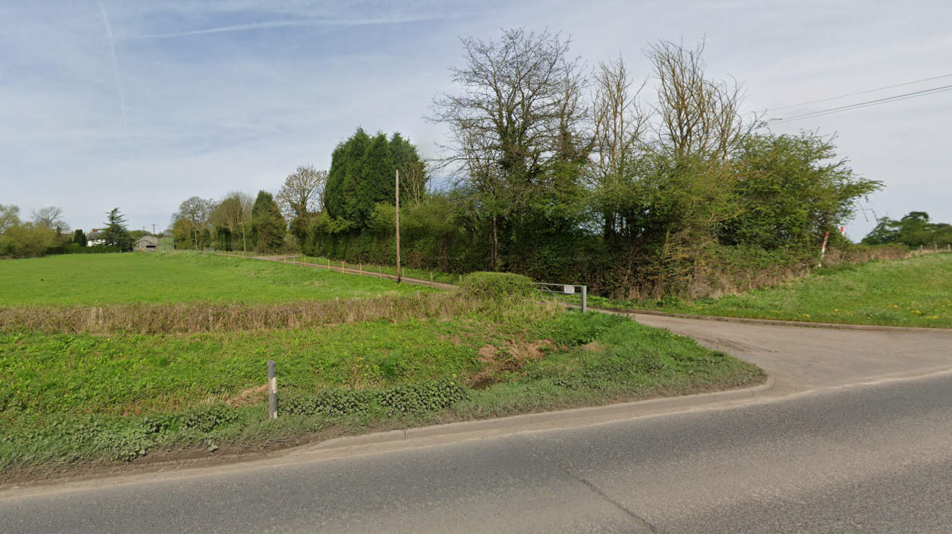 A Google Streetview of farmland off a main road. A track runs between a green field on one side and trees on the other to a farm site in the distance.