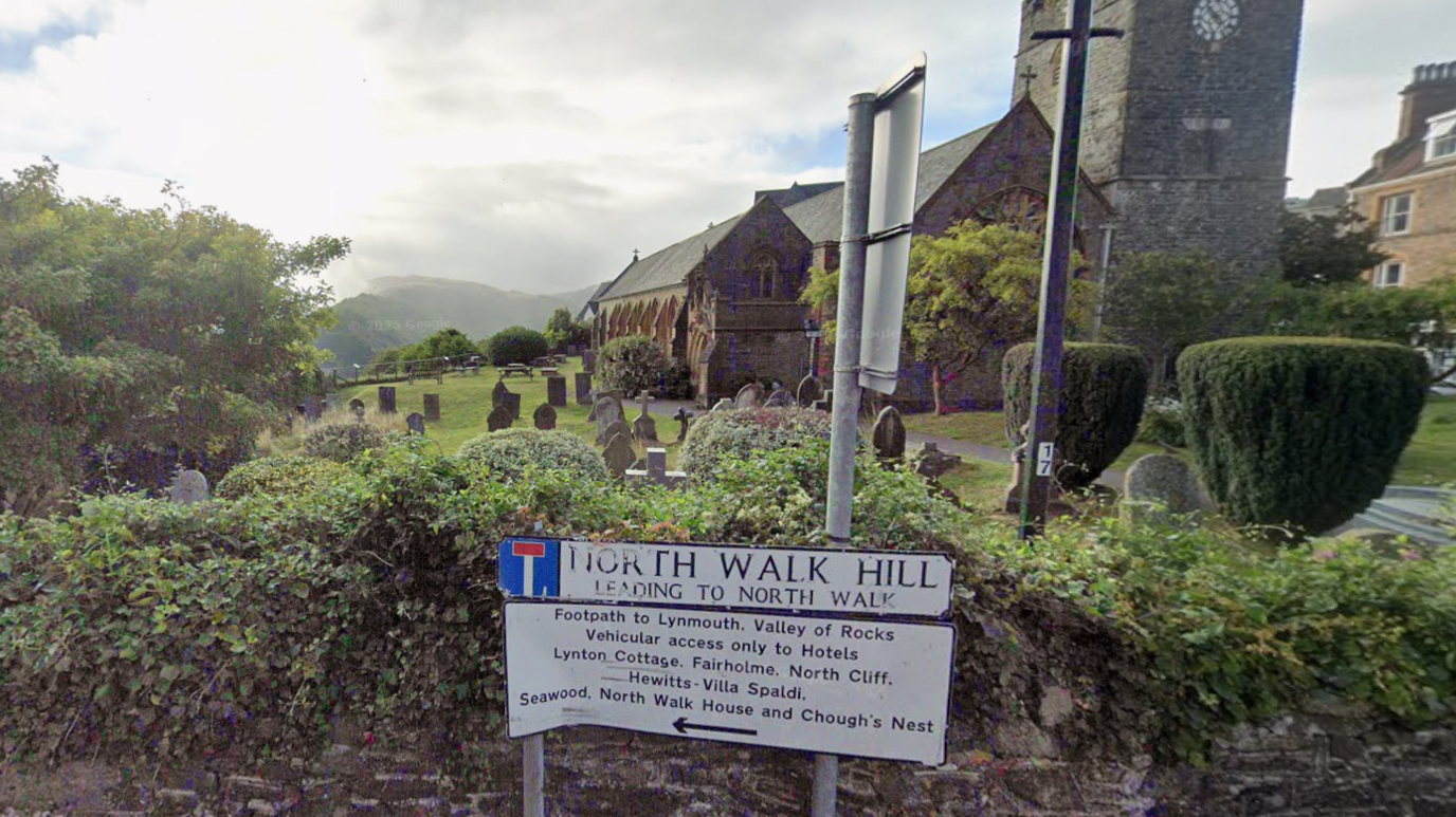 A signpost reads: North Walk Hill leading to North Walk and underneath a footpath to Valley of Rocks is detailed along with what areas the road offers to vehicles. There is a stone wall behind the sign which leads on to a churchyard with yew trees, gravestones and a church. There are hills in the distance. 