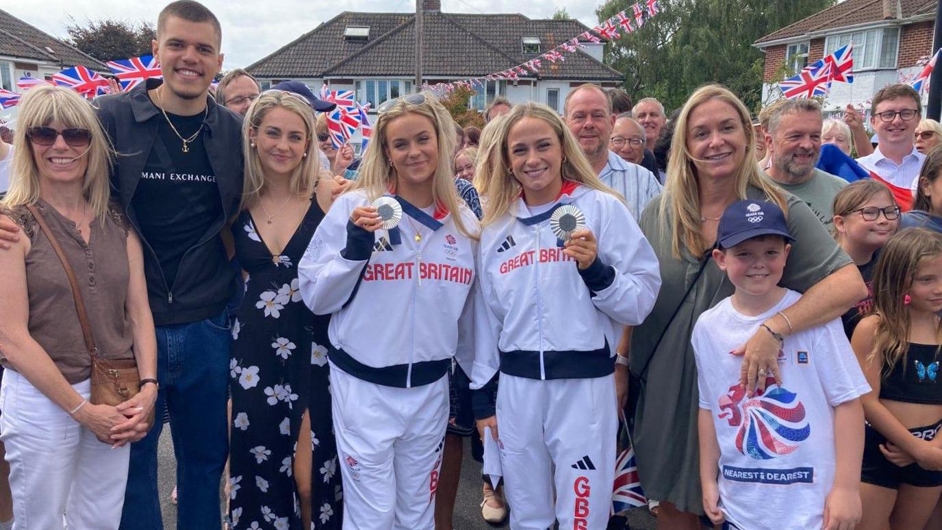 Kate and Isabelle wearing Team GB tracksuits wearing their silver medals, surronded by their family, friends and neighbours, outside on their cul-de-sac