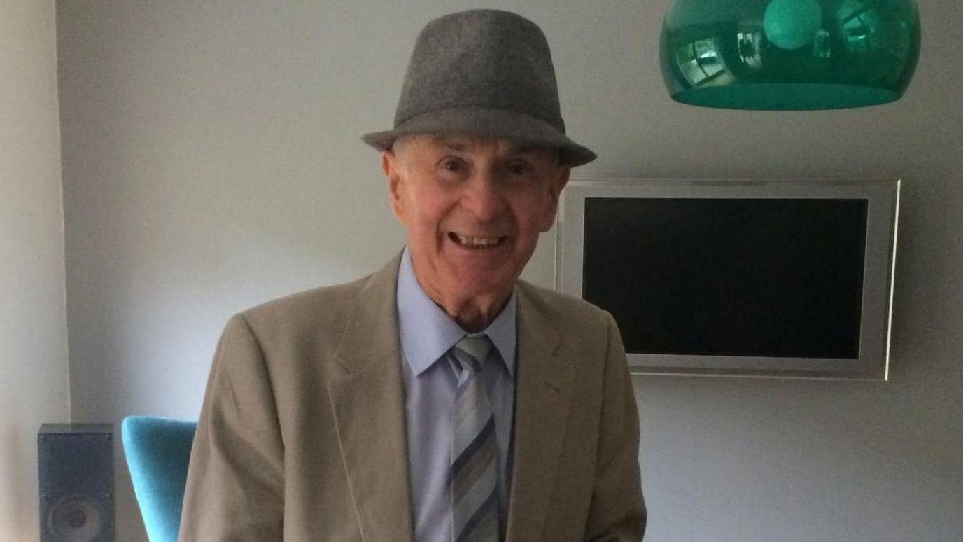 An elderly man smiles for a photo. He is wearing a grey hat and a cream jacket and light blue shirt.