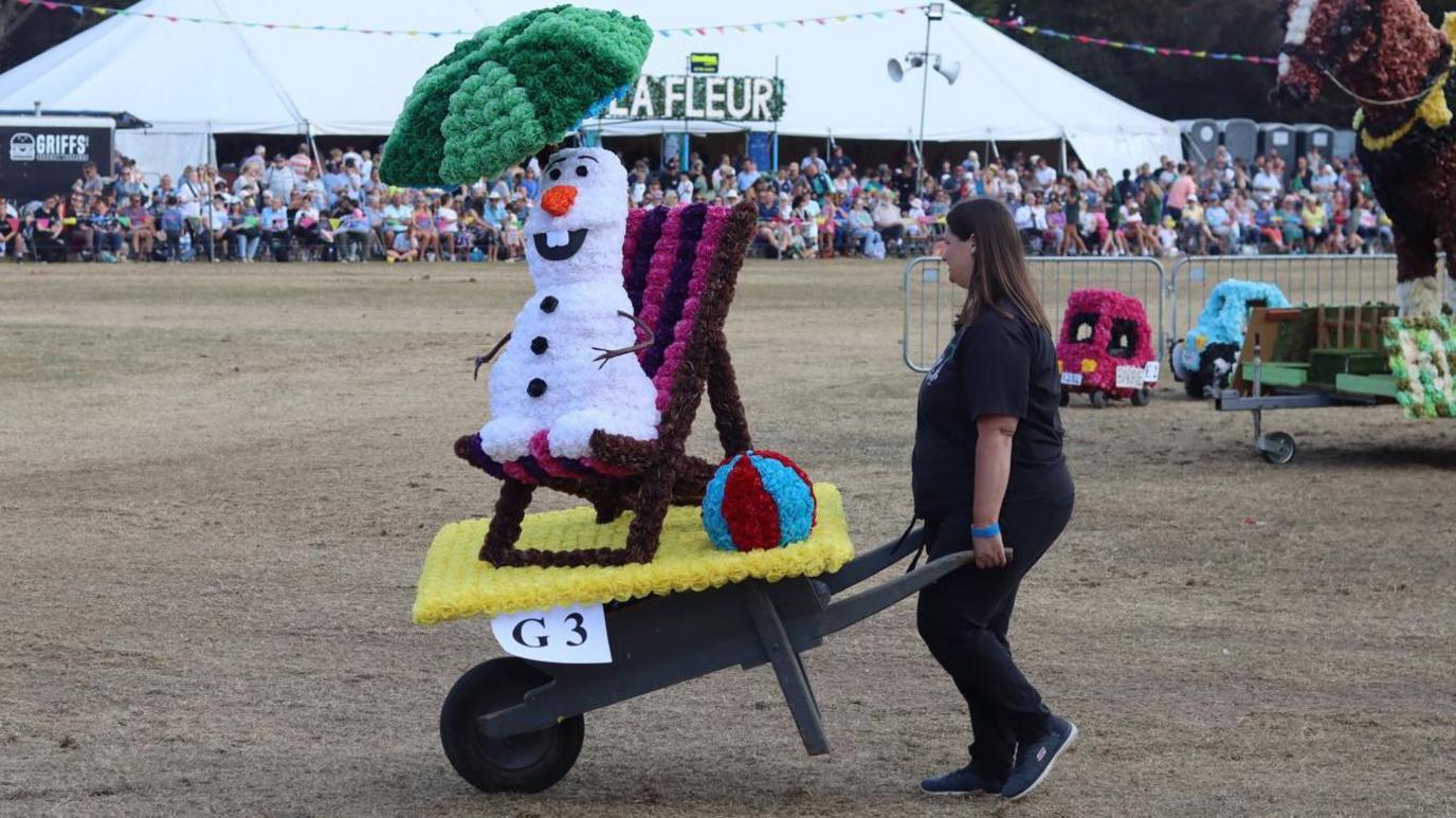 A woman pushes a wheelbarrow along the showground during the North Show's Battle of Flowers in Guernsey. Her design features Olaf from Frozen sitting on a deckchair.