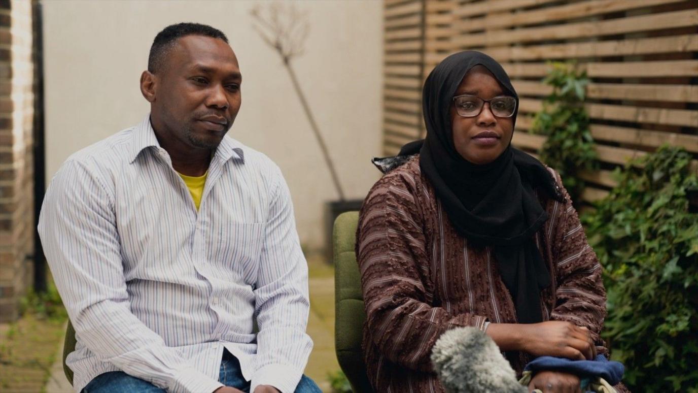 Mr Mohammed is sat on the left, wearing a striped shirt with a yellow top underneath and Mrs Mohammed, who is sat next to her husband, is wearing glasses, a hijab and a long red top.