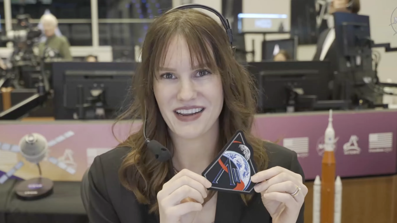 Leah Cheshier Mustachio is wearing a smart suit and has long brown hair. She is sat in mission control. She is holding up the patch to the camera. You can see the earth in the foreground and the moon behind it