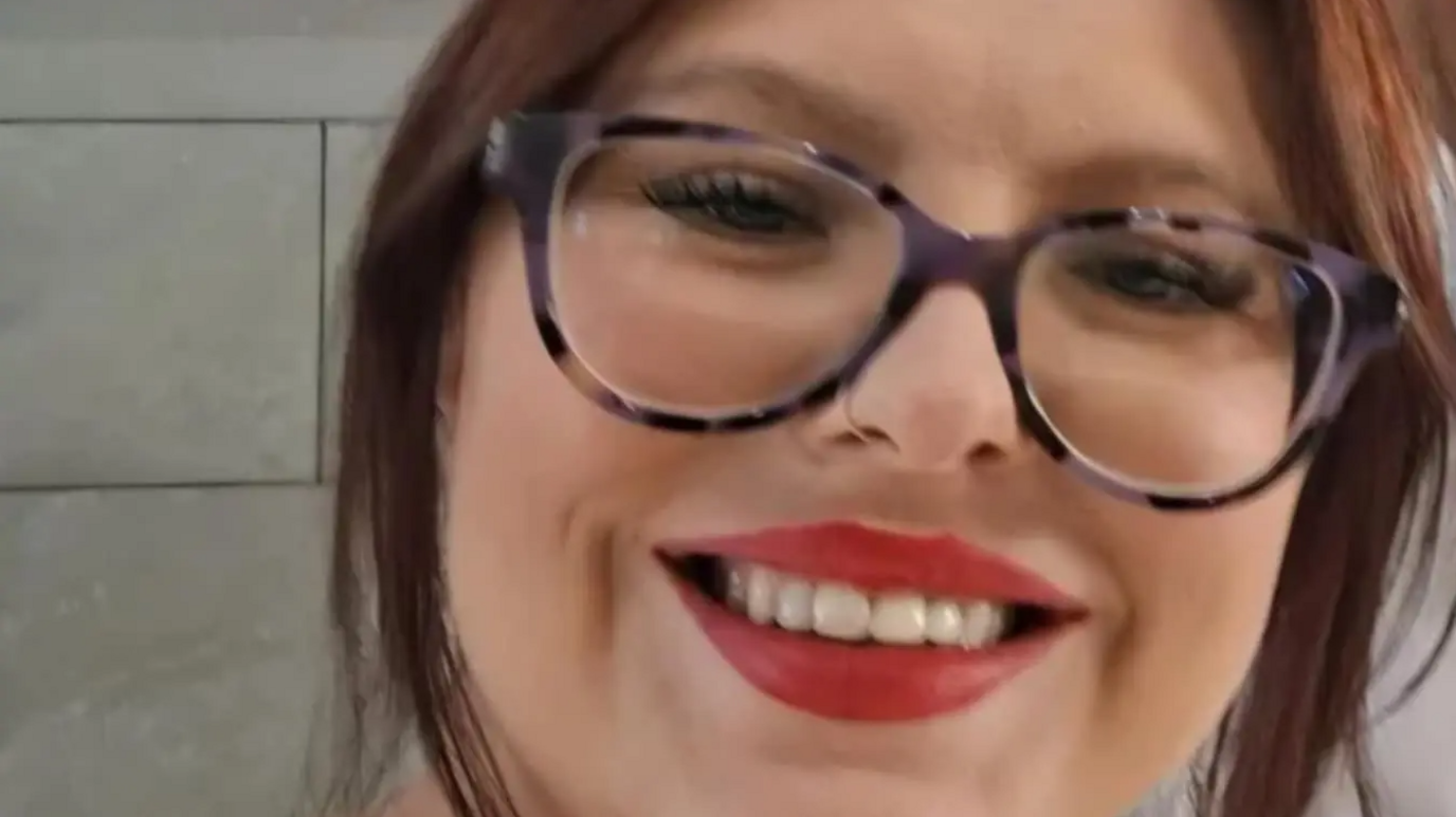 Cassie Marriot has dark brown hair and is wearing purple and black framed glasses. She is wearing red lipstick. 