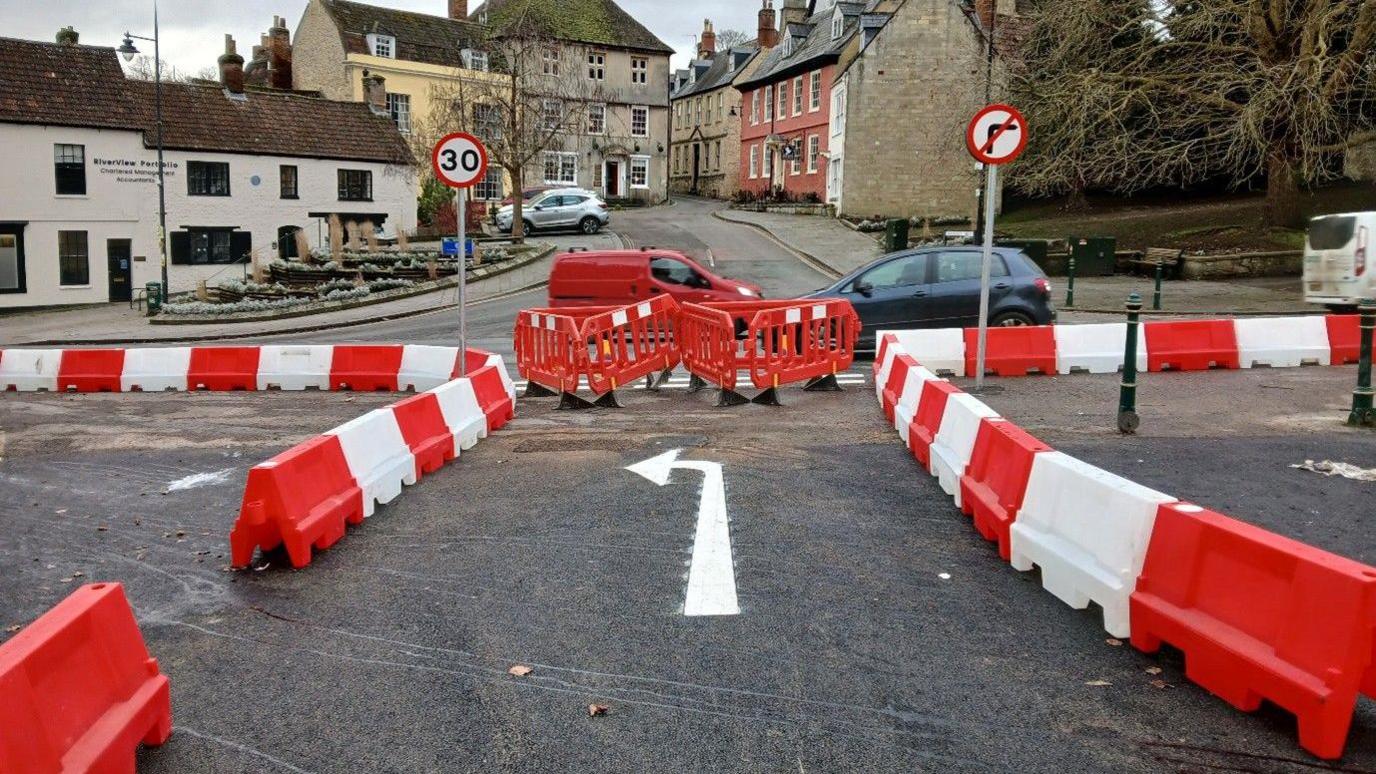 The junction of a one-way street with a 'no right hand turn' sign and 30mph speed limit sign at the end of it. Plastic red and white barriers line the edge of a road, creating a barrier between cars and pedestrians. The road is closed at the end by red bollards.