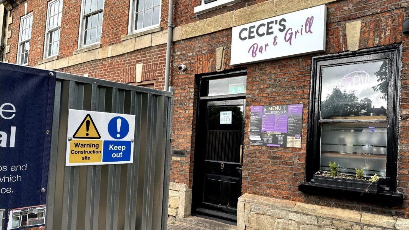 An old brick building with a sign reading 'Bar and Grill', and a menu outside. In the street there are hoardings with construction-site warning signs