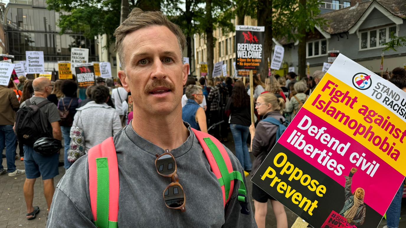 Luke Philpott has a moustache, short brown hair, sunglasses folded into his grey shirt and a pink and green rucksack. He's also holding a sign that says: Stand up to racism. Fight against Islamophobia, defend civil liberties, oppose Prevent